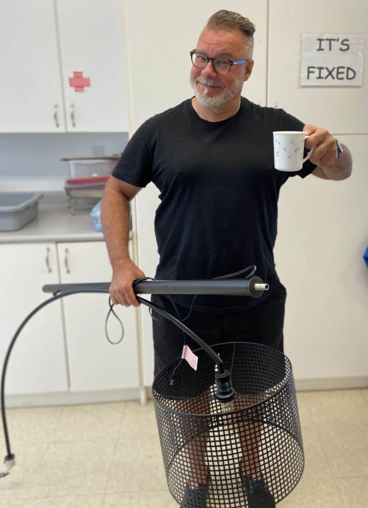 Photo of a smiling man holding a lamp in front of a sign that says "It's Fixed".